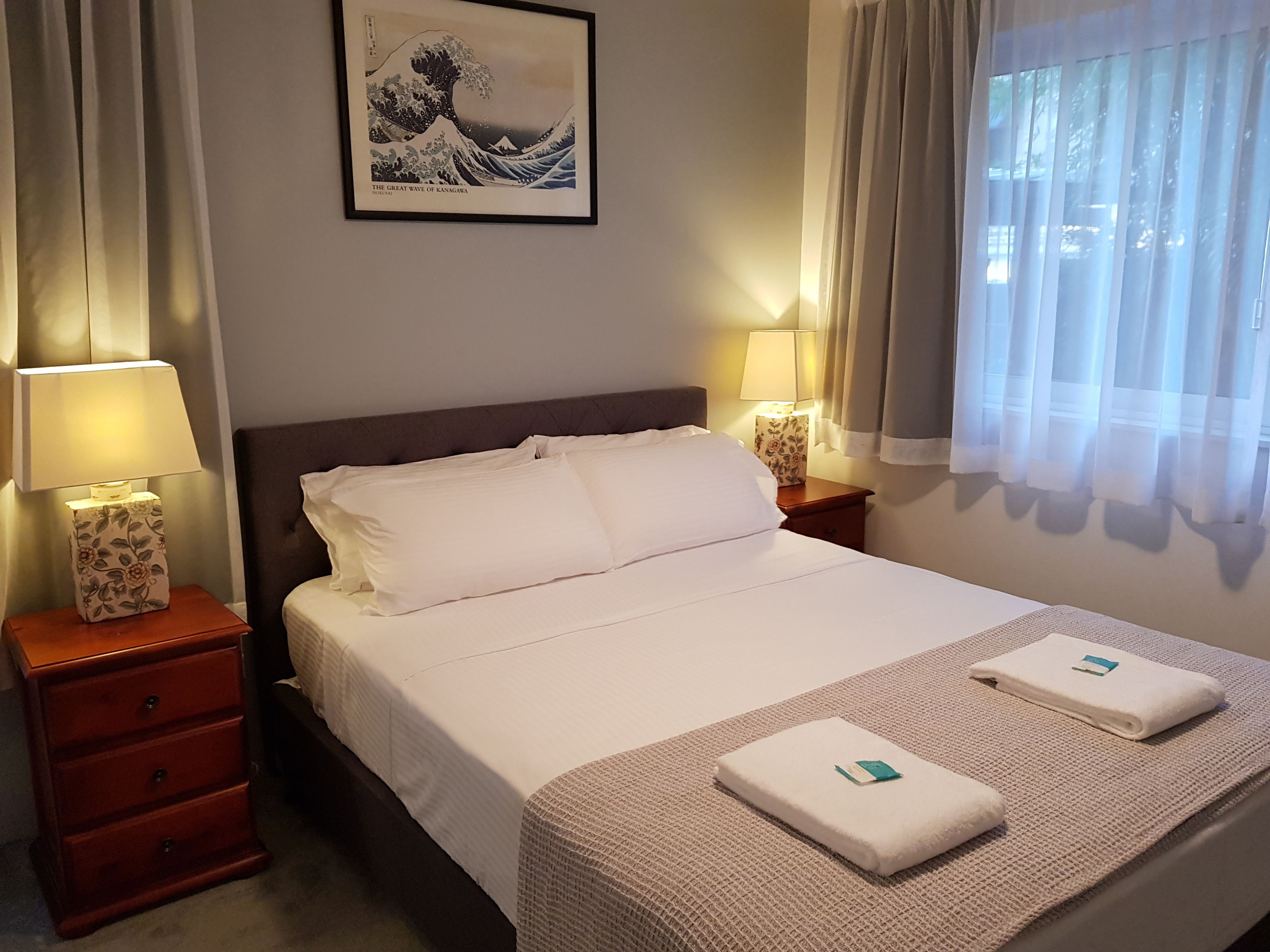 Un lit bien fait, avec du linge de lit blanc, deux tables de chevet avec des lampes, une œuvre d’art encadrée au mur et une fenêtre avec des rideaux légers.
