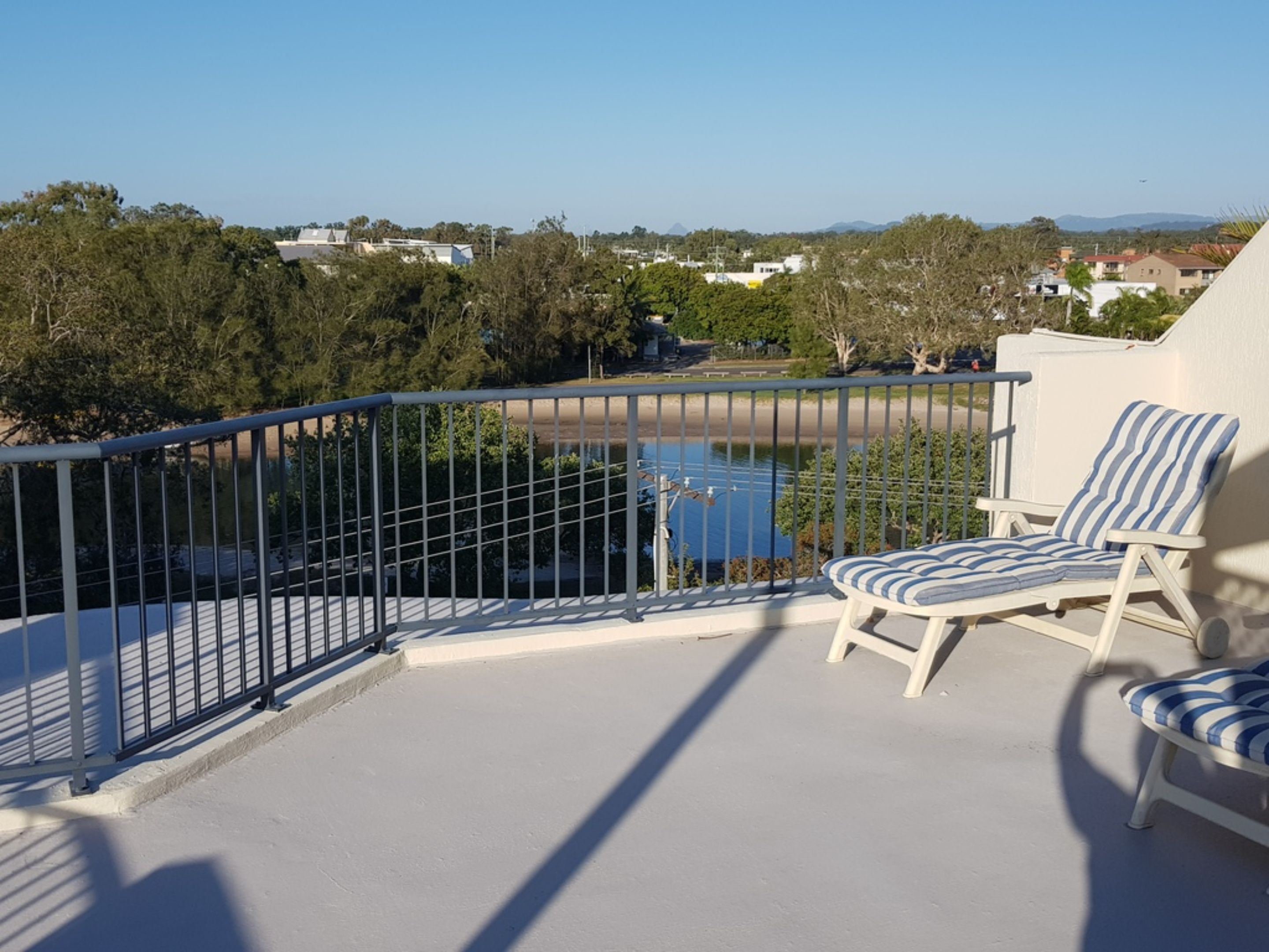 Un balcón con barandilla de metal, dos sillones a rayas y vista a un cuerpo de agua y edificios distantes.
