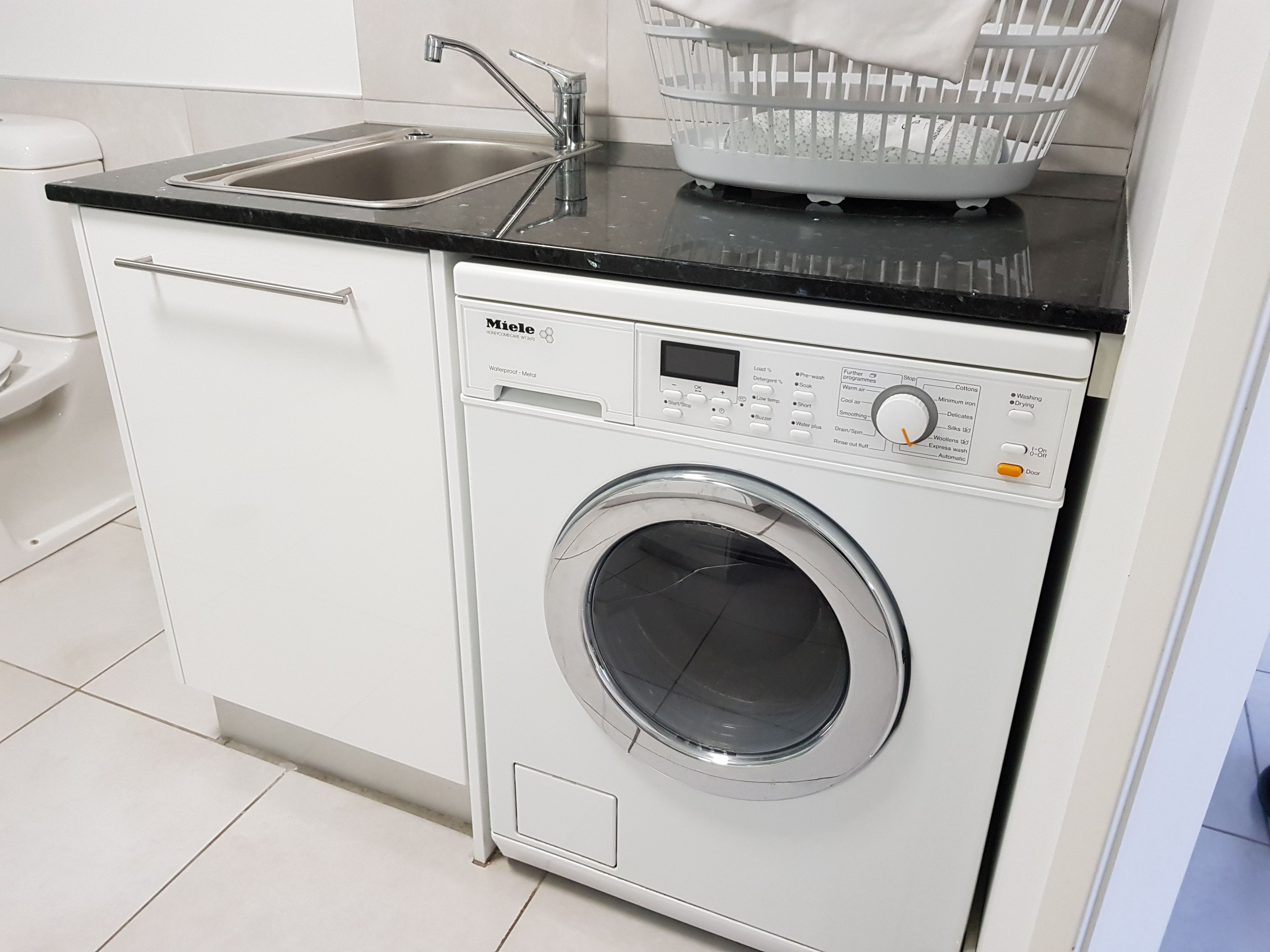 Une buanderie moderne et compacte, dotée d’un lave-linge et d’un sèche-linge encastrés, d’un évier et d’un panier posé sur le plan de travail.