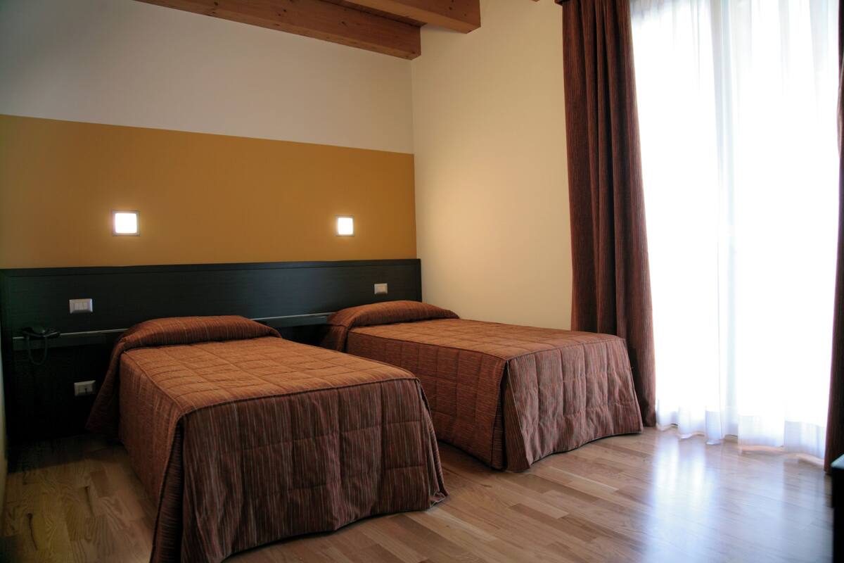 A room with two single beds, a wooden ceiling, a nightstand with a lamp, and a window with curtains.