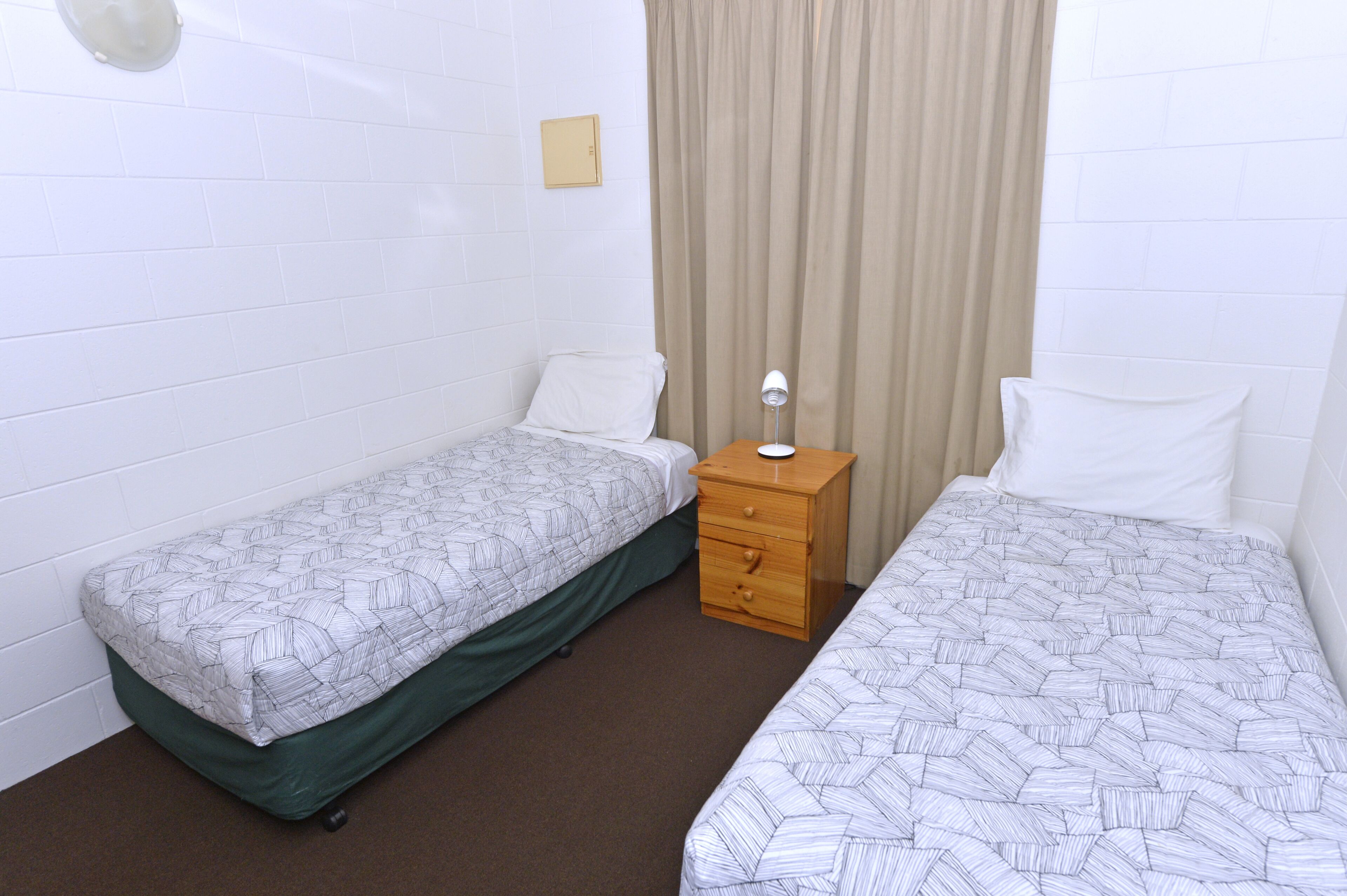 A small, clean room with two single beds, a wooden nightstand, and a beige curtain.