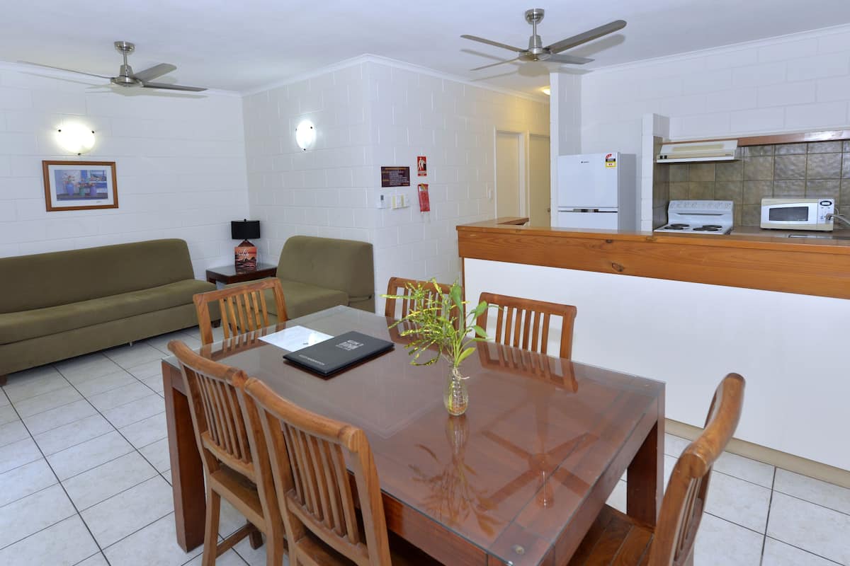 Un comedor con una mesa de madera, sillas y un florero con una planta. Hay una cocina con estufa, microondas y refrigerador.