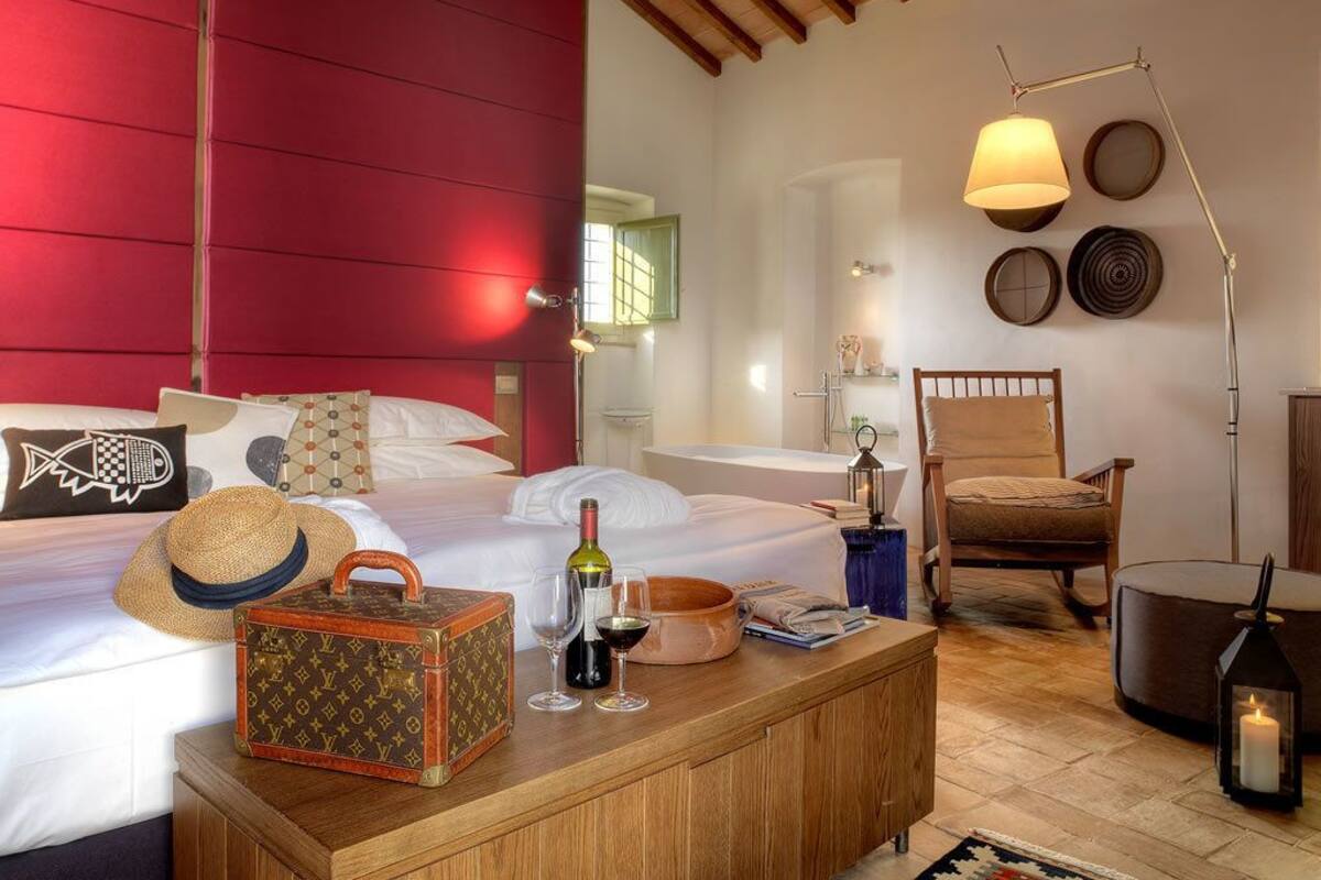 Un dormitorio con cabecera roja, mesita de noche de madera con una maleta, copas de vino y un sombrero. Hay una silla y una lámpara de pie en la habitación.