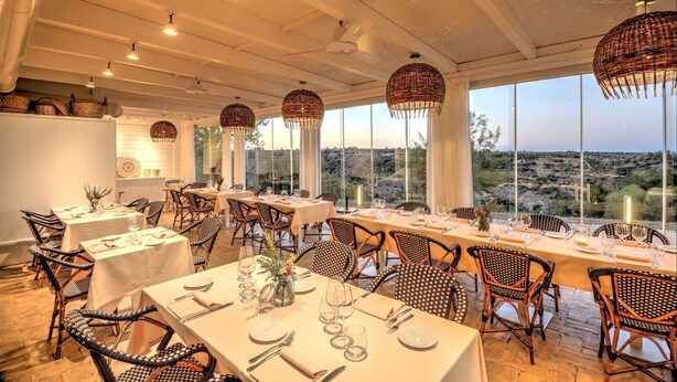 A restaurant with multiple tables set for dining, wicker chairs, and large windows offering a view of the landscape. - Torre Fiore Hotel Masseria (Pisticci)