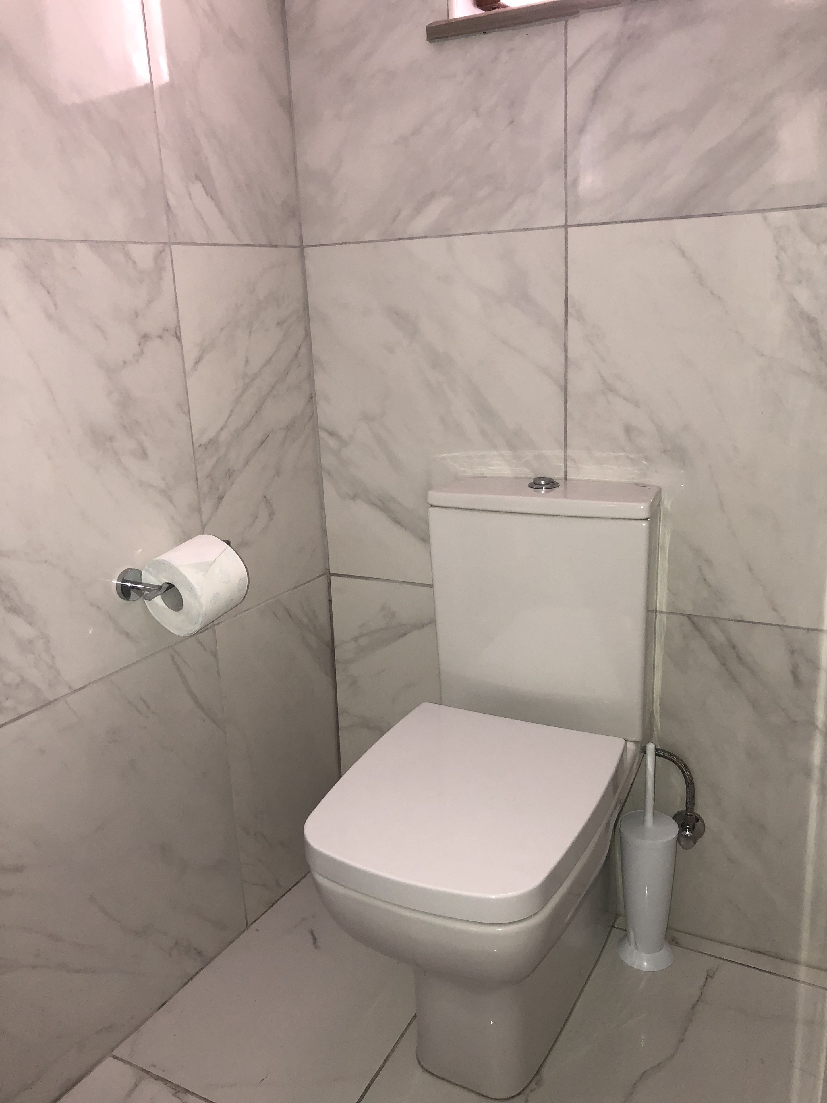 Un baño con inodoro, azulejos de mármol, dispensador de papel higiénico y cabezal de ducha.