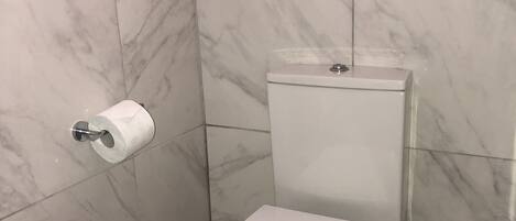 Un baño con inodoro, azulejos de mármol, dispensador de papel higiénico y cabezal de ducha.