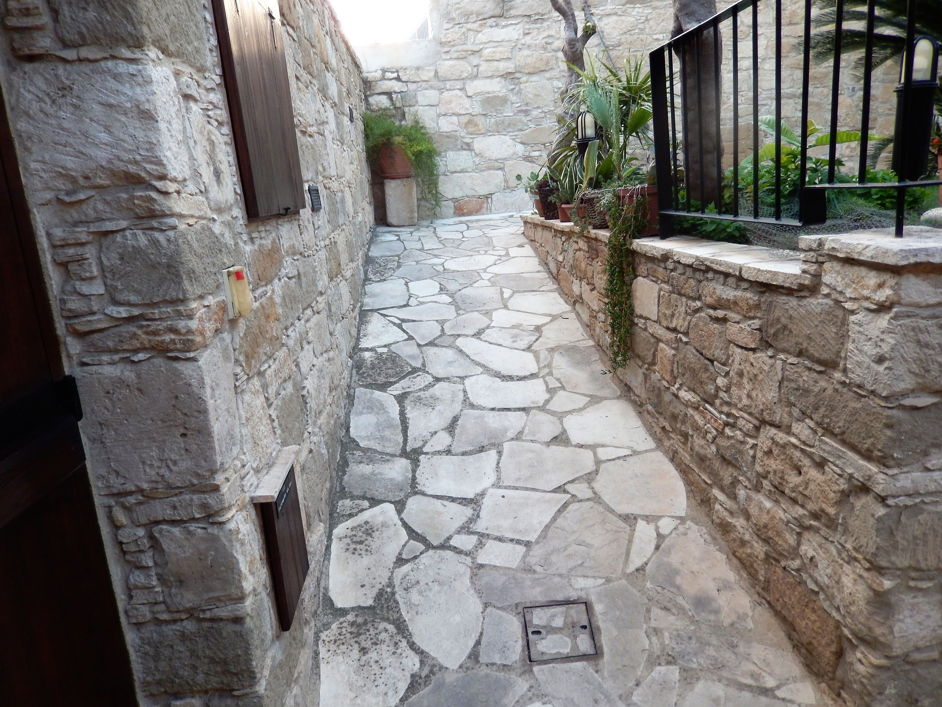 A narrow stone-paved alleyway with stone walls, a metal gate, and potted plants.