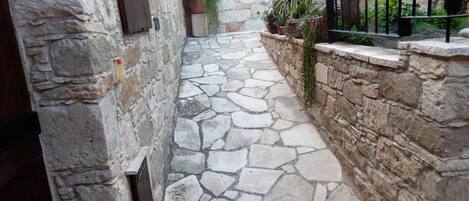 A narrow stone-paved alleyway with stone walls, a metal gate, and potted plants.
