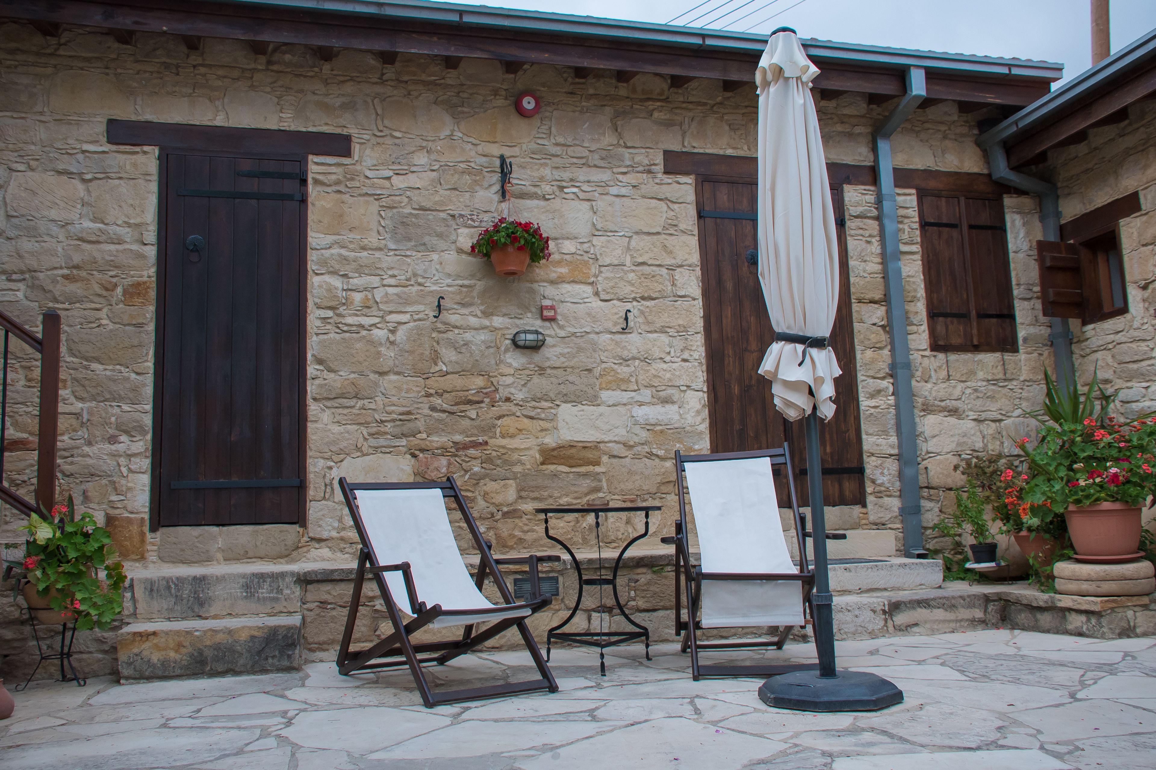 A stone building with a wooden door and shutter, outdoor seating with a table and chairs, and potted plants.