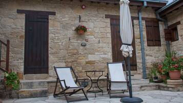 A stone building with a wooden door and shutter, outdoor seating with a table and chairs, and potted plants.