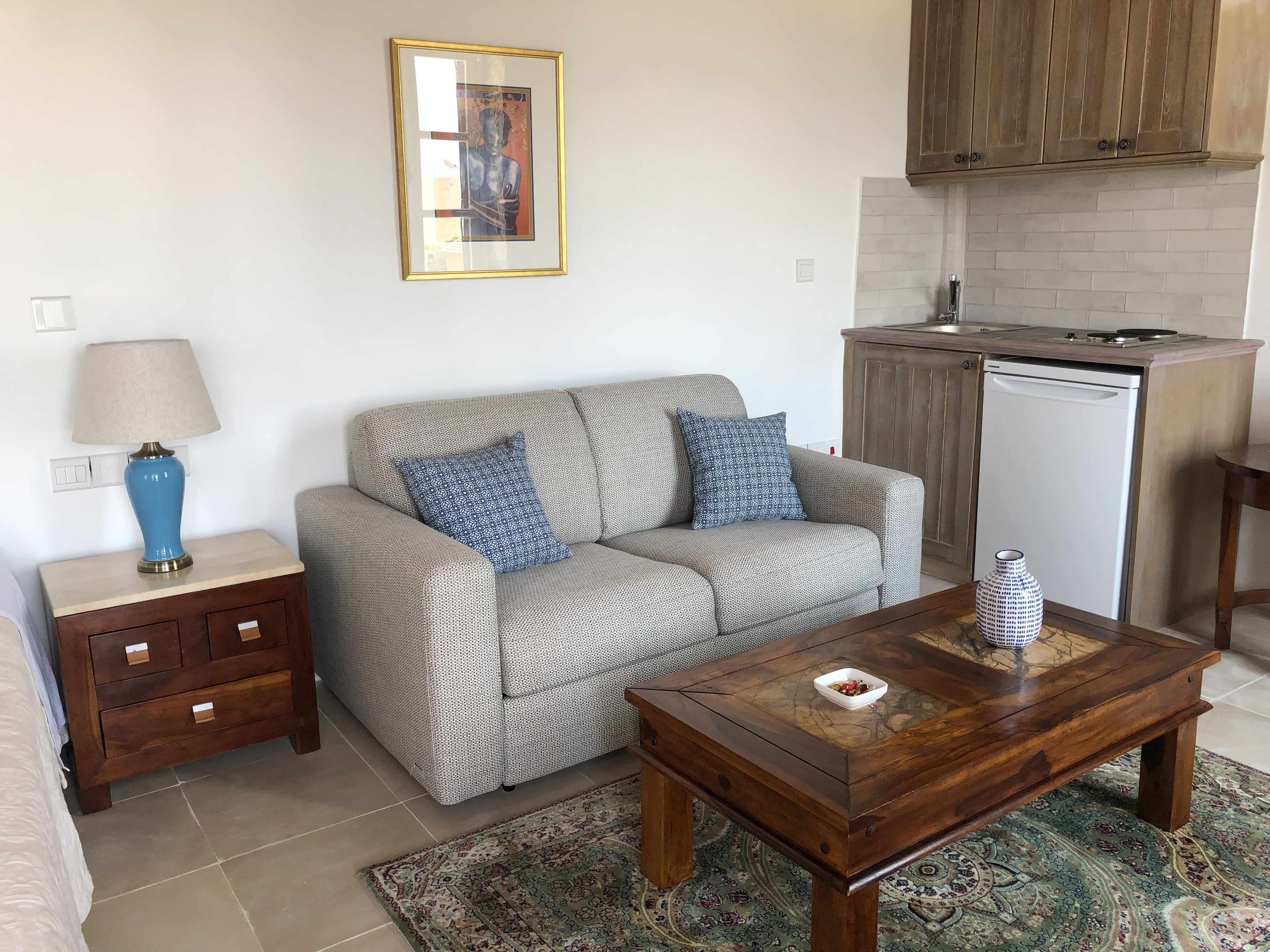 A living room with a sofa, a coffee table, a lamp, a rug, and a kitchen area with a dishwasher.