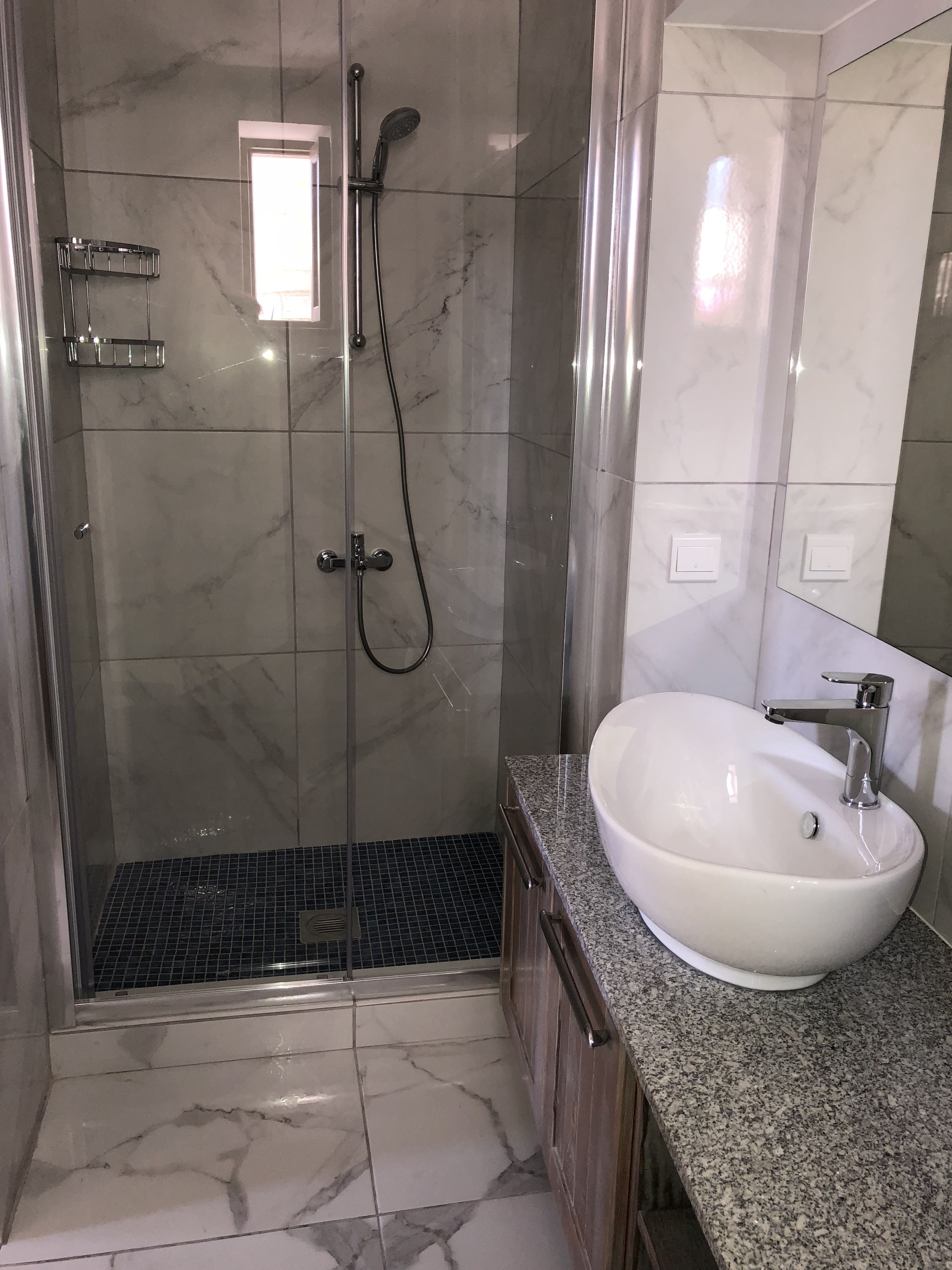 A modern bathroom with a glass-enclosed shower, a white sink, and marble flooring.