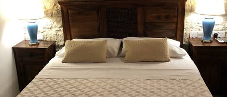 A bed with a patterned bedspread, two pillows, a wooden headboard, and a framed artwork above the bed. There are two bedside tables with lamps on either side of the bed.