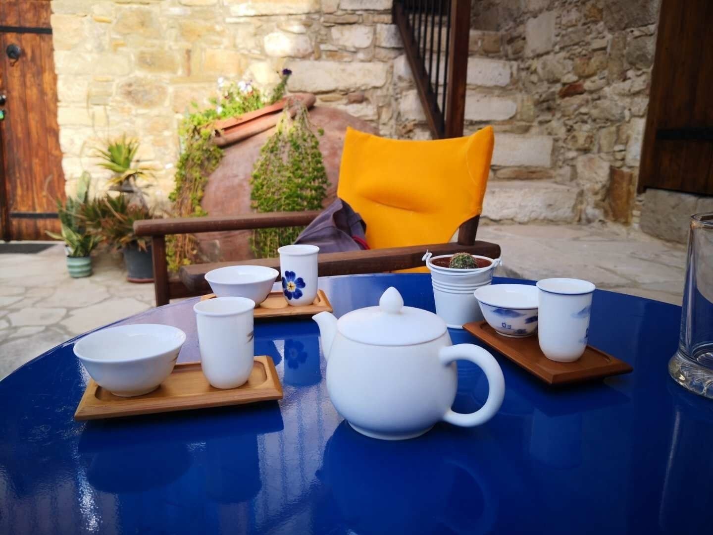 A table set with white teacups, a teapot, and a blue tablecloth in a stone-walled courtyard with potted plants and a wooden chair.