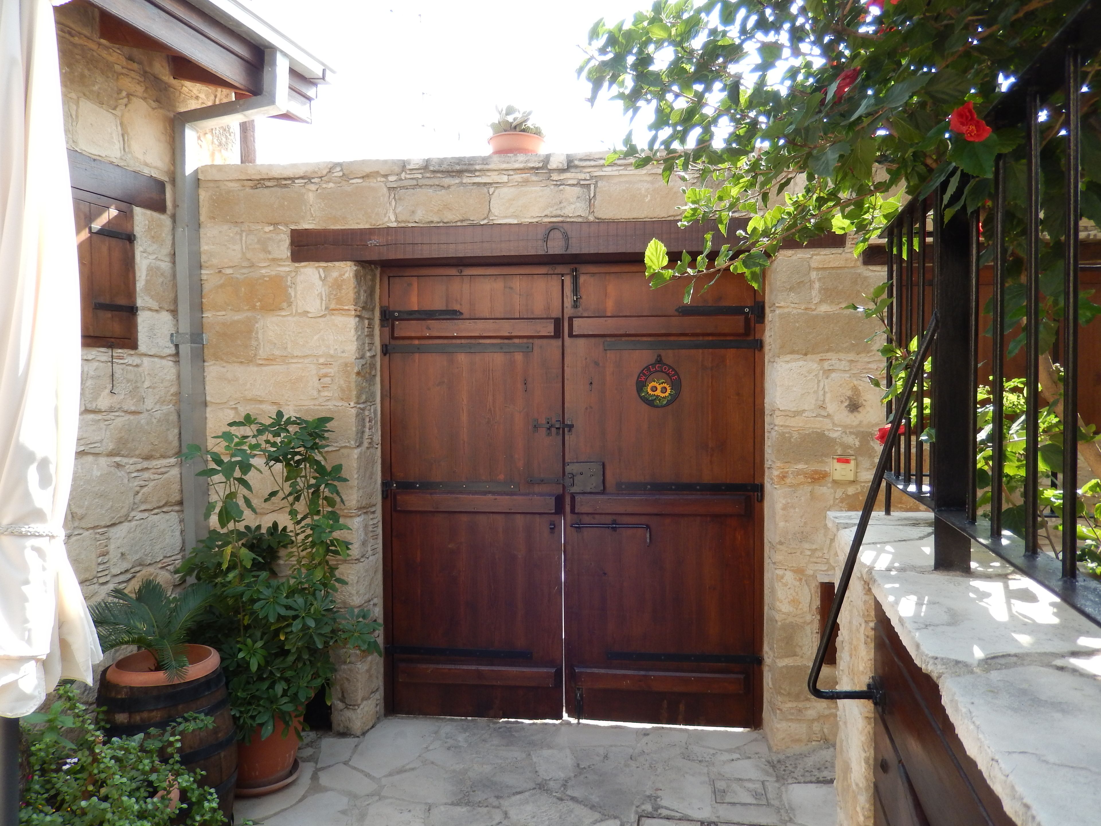 Puerta doble de madera con un letrero que dice 'Welcome', ubicada en una pared de piedra con plantas en macetas.