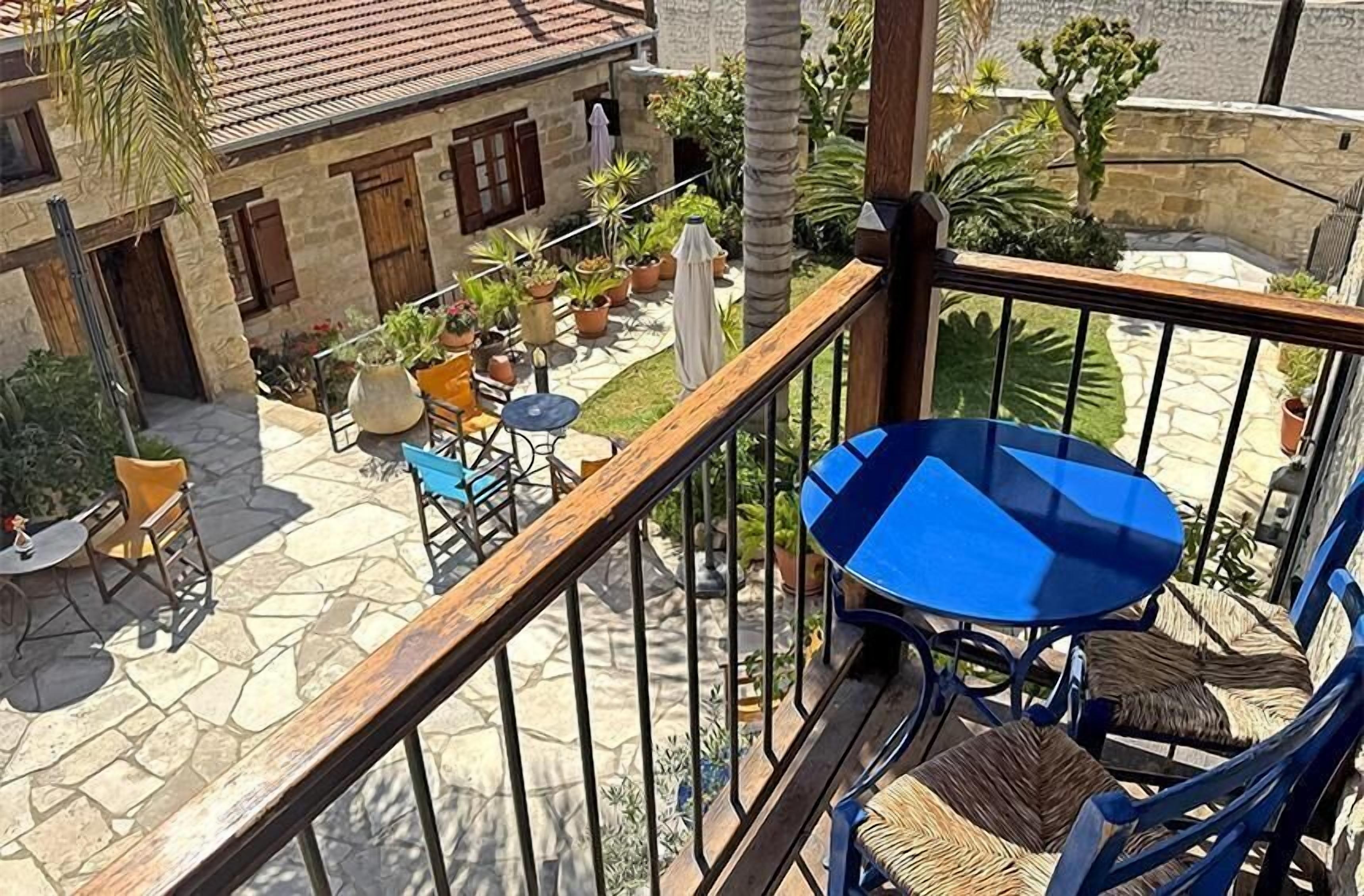 A balcony with a blue table and wicker chairs, overlooking a courtyard with stone flooring, potted plants, and a building with wooden shutters.