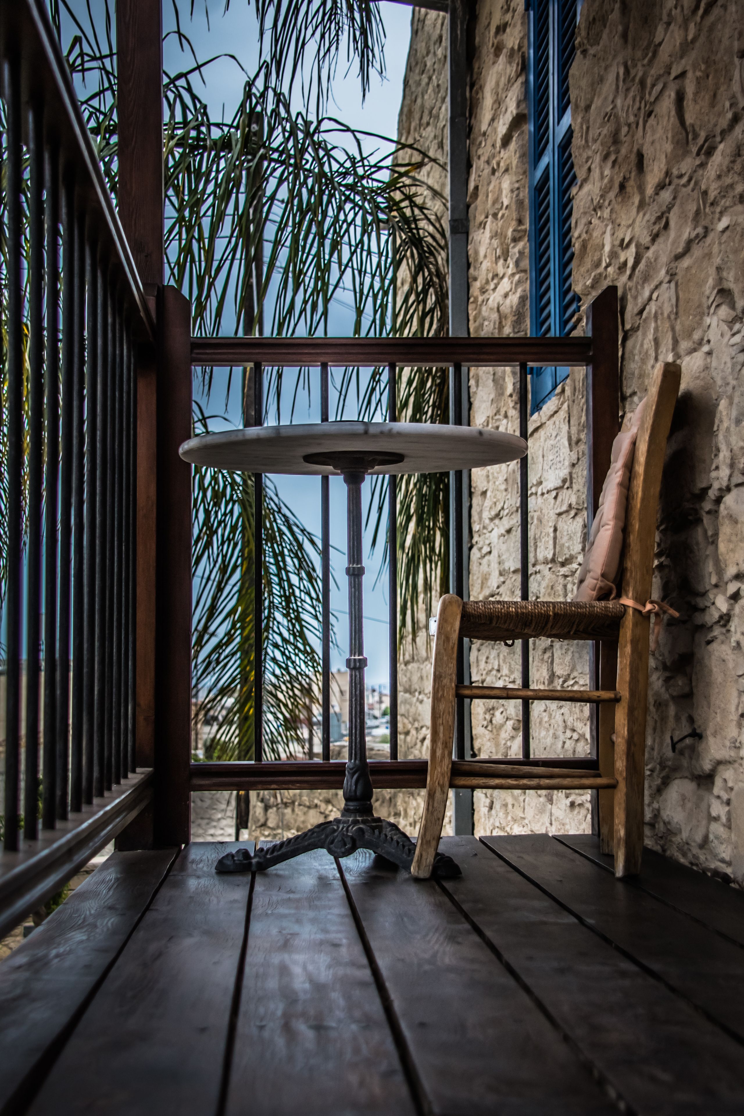 Una mesa y silla de madera en un balcón, con una pared de piedra y contraventanas azules al fondo.