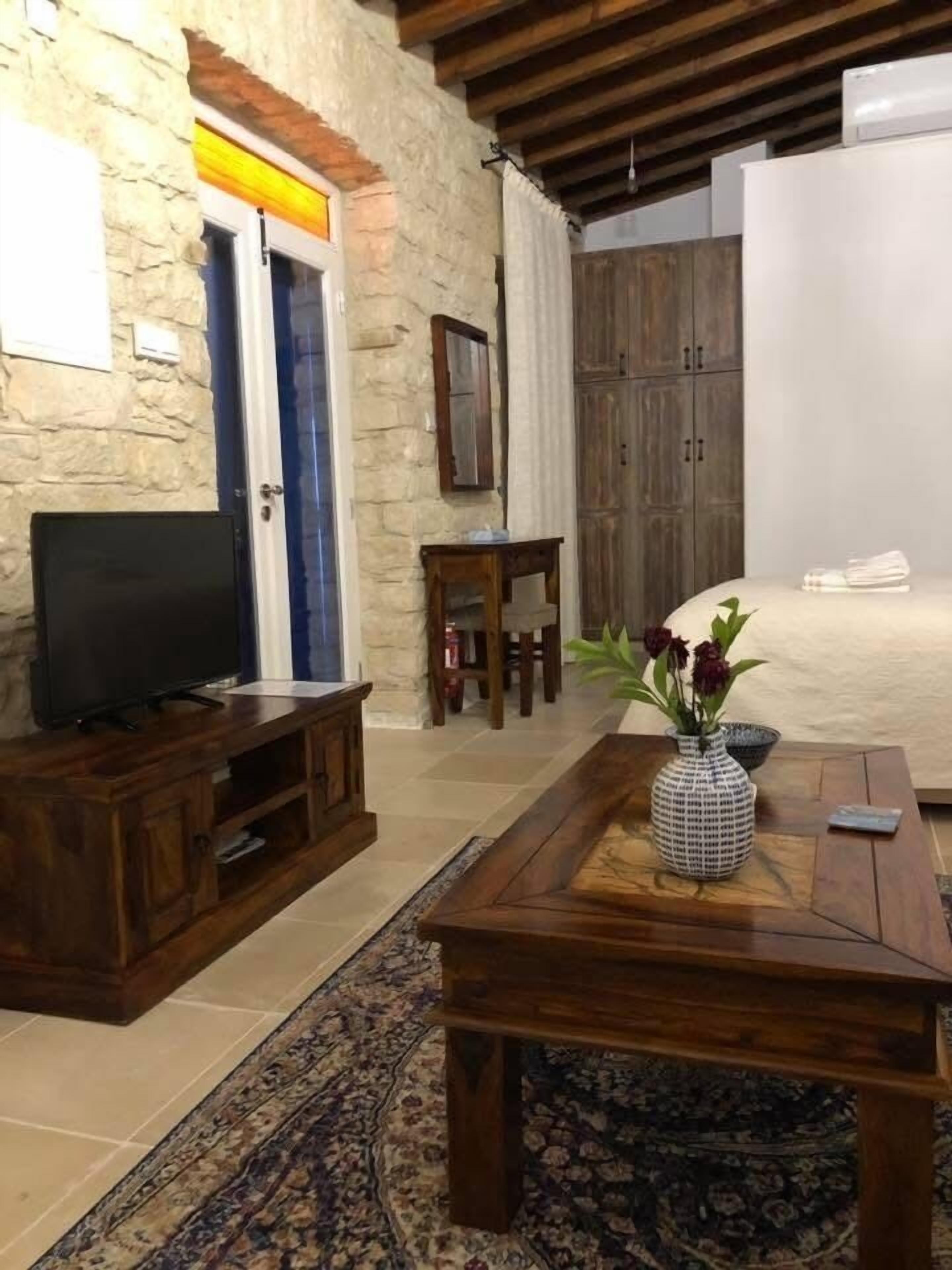 Una sala de estar con pared de piedra, muebles de madera, un televisor, una mesa de centro con un jarrón y una cama con cabecero blanco.