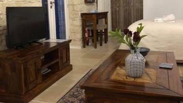 Una sala de estar con pared de piedra, muebles de madera, un televisor, una mesa de centro con un jarrón y una cama con cabecero blanco.