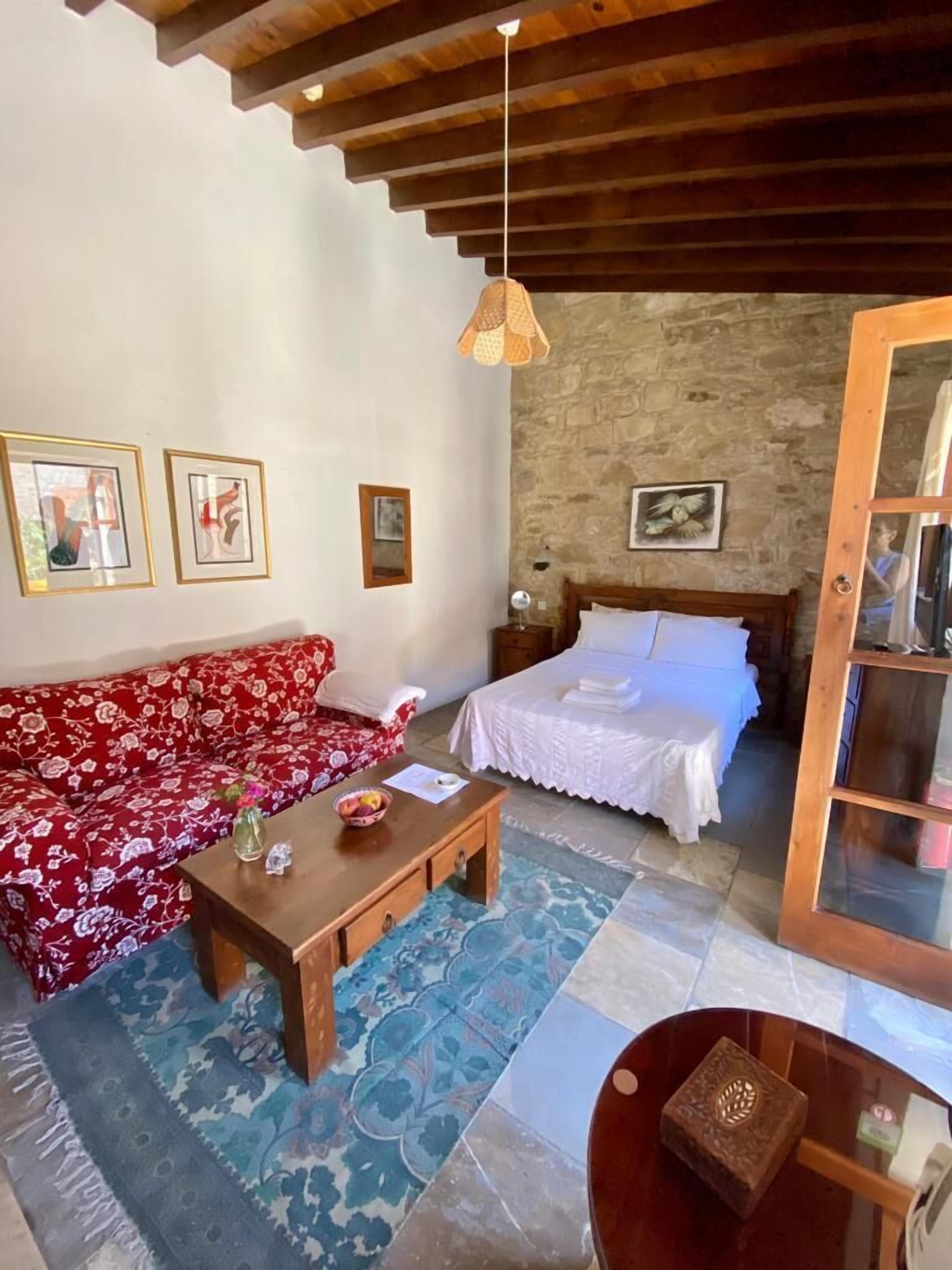Une pièce chaleureuse avec un canapé rouge à motifs floraux, une table basse en bois, un lit recouvert de draps blancs et un mur en pierre.