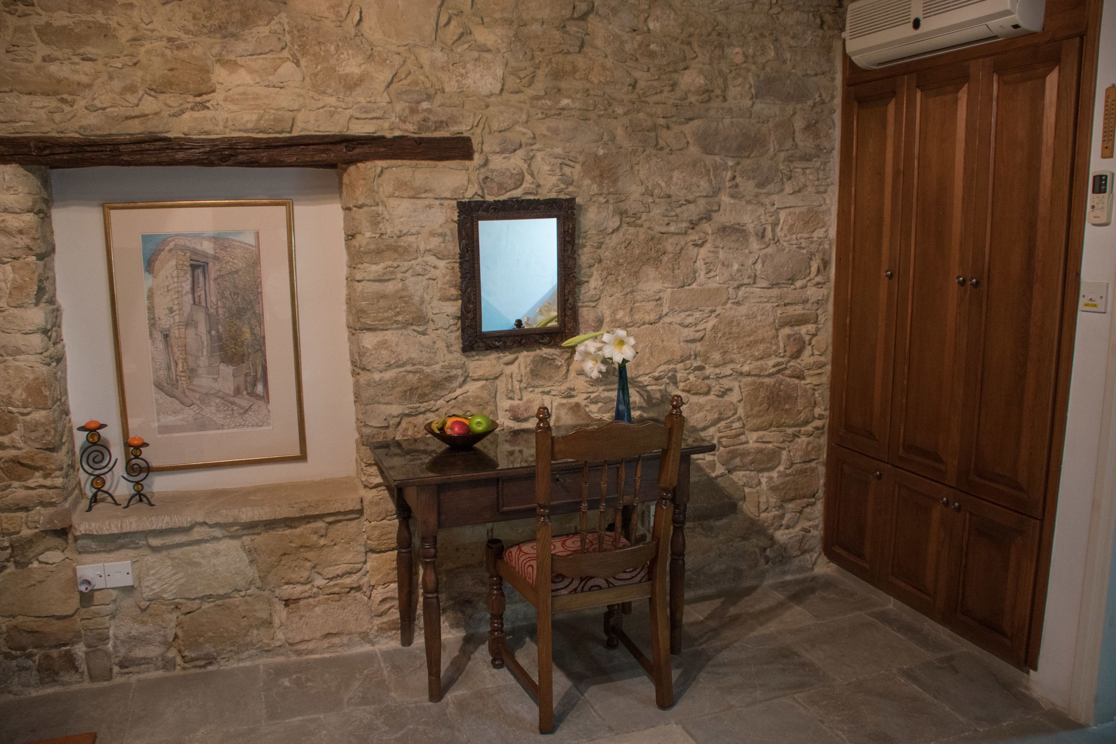 A stone wall with a framed picture, a mirror, a wooden chair, a small table with a fruit bowl, and a wooden wardrobe.