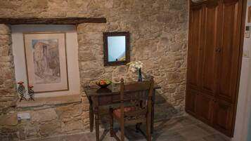 A stone wall with a framed picture, a mirror, a wooden chair, a small table with a fruit bowl, and a wooden wardrobe.