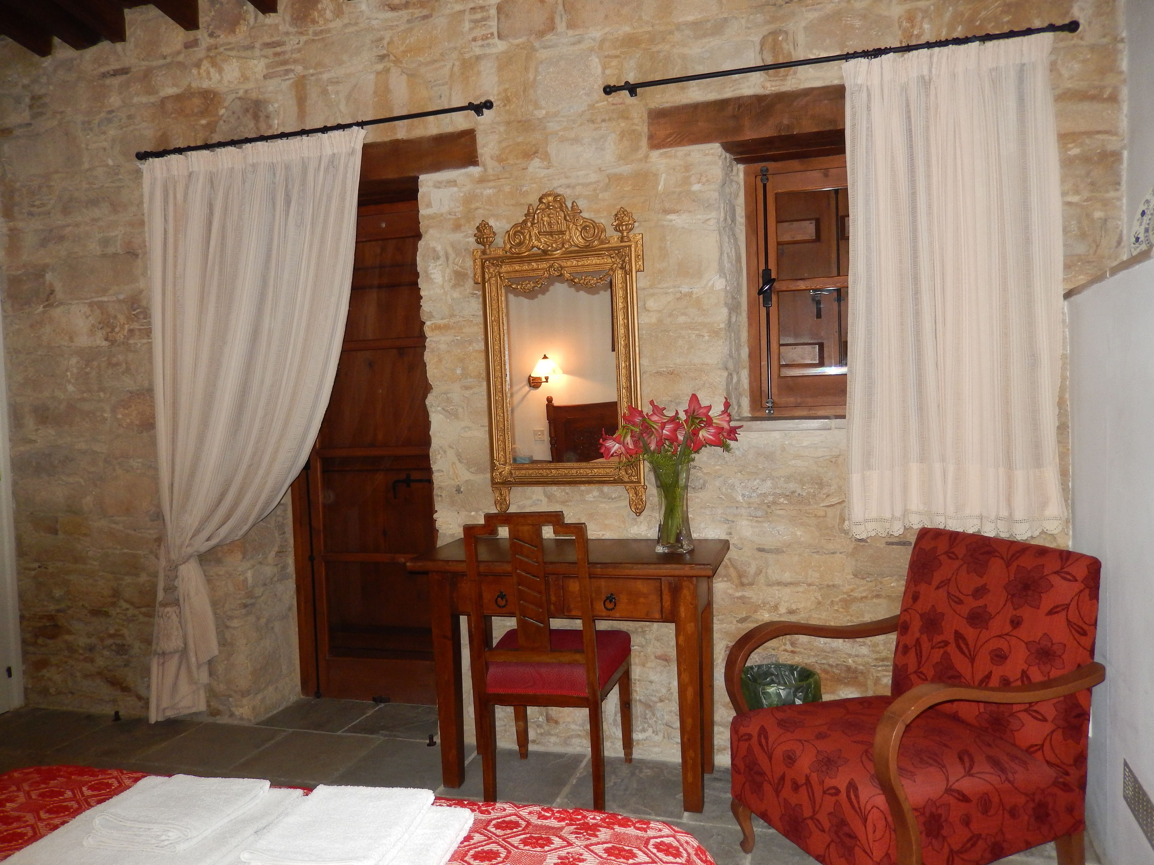 Una habitación con paredes de piedra, una cama de madera, un escritorio de madera con una silla, un sillón rojo con motivos florales, un espejo y un jarrón con flores.