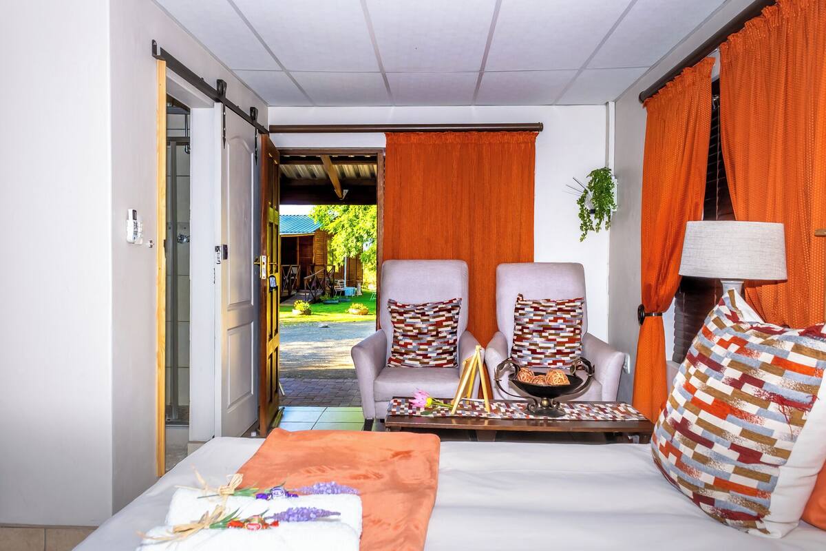A hotel room with a bed, two armchairs, a table with a tray of snacks, and orange curtains.
