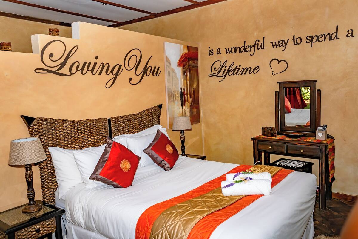A neatly made bed with a red and gold bedspread, a wall with a 'Loving You' sign, and a mirror on a wooden nightstand.