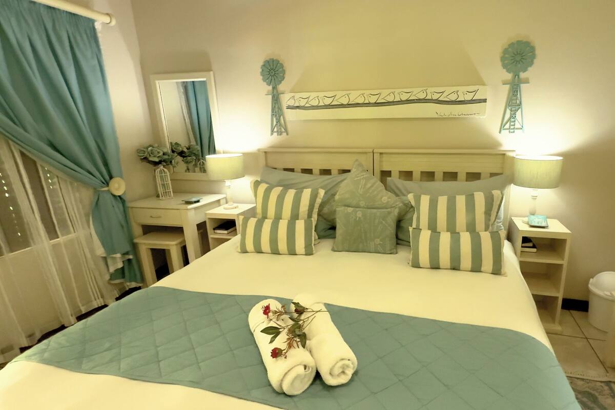 A neatly made bed with striped pillows, a quilted blue bedspread, and rolled towels with flowers.