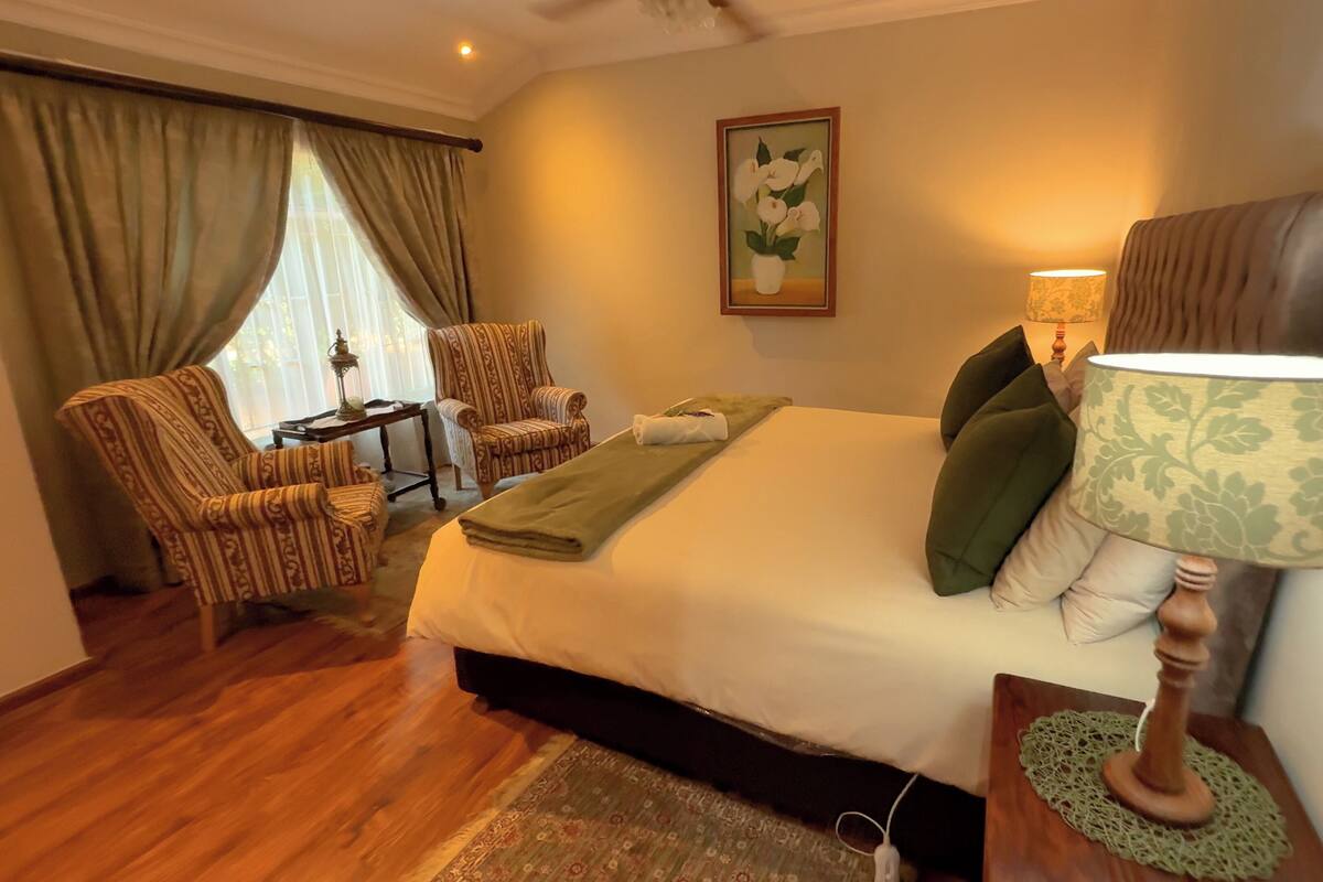 A hotel room with a bed, two chairs, a table with a lamp, and a painting on the wall.