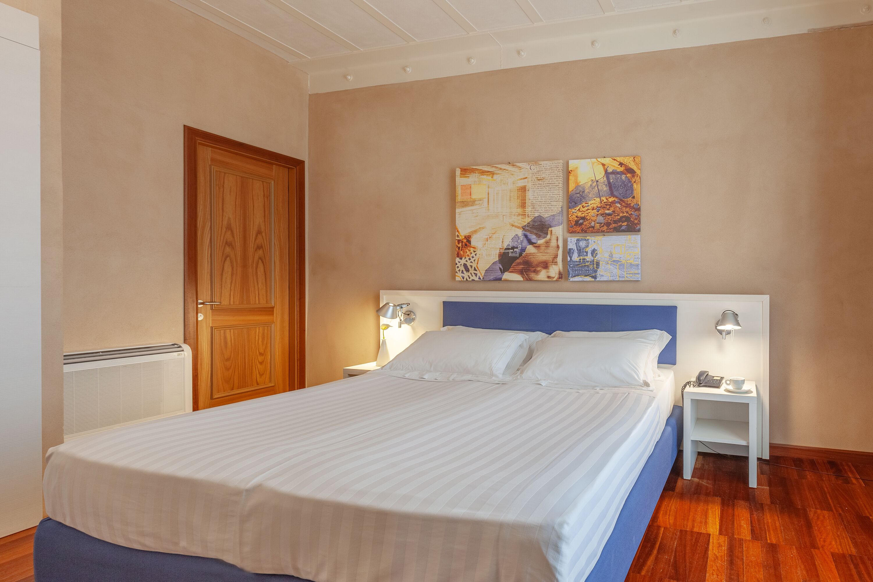 Un letto rifatto con cura, con una testiera blu, due comodini con lampade e una parete decorata con arte astratta.