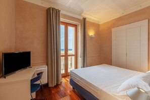 A hotel room with a bed, a TV, a desk with a chair, a radiator, and a view of the sea through a window.