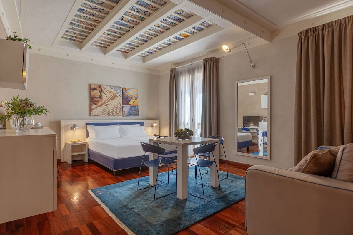 Una habitación de hotel moderna con cama, una mesa de comedor con sillas, un sofá, un espejo grande y un techo con baldosas estampadas.