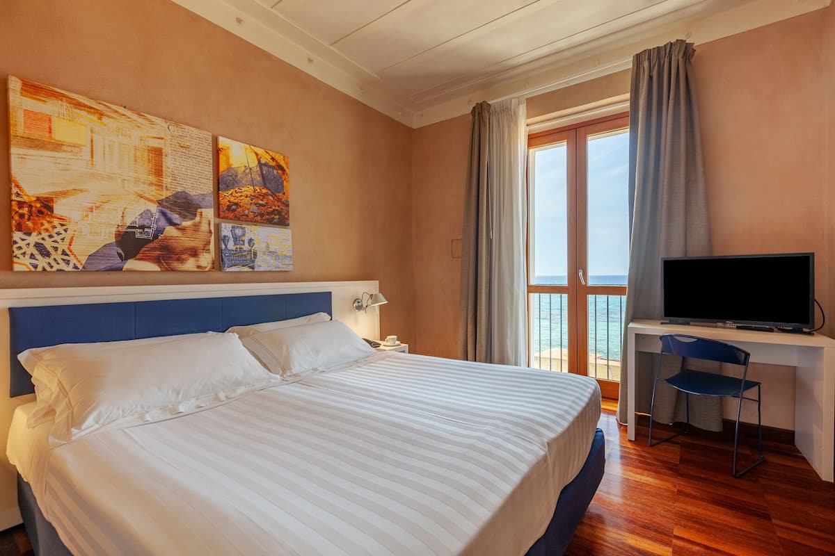 Habitación de hotel con una cama grande, un escritorio con televisor y vista al mar desde la ventana.