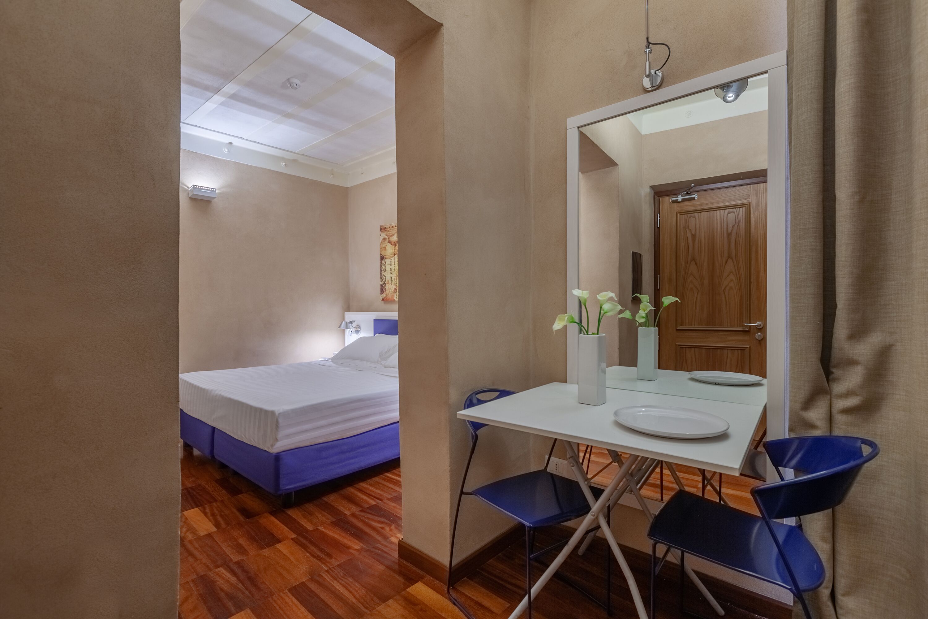 A compact room with a bed, a small table with chairs, a mirror, and a door.