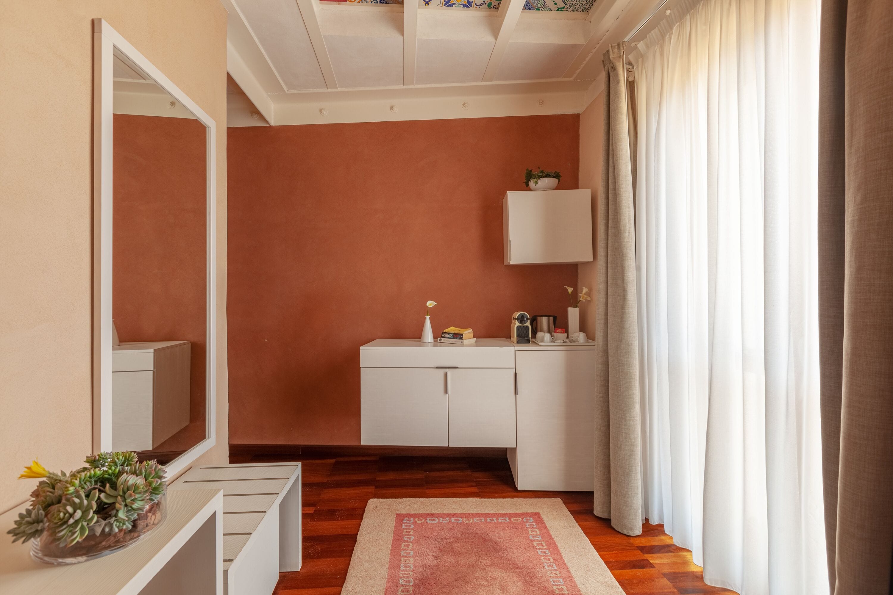Una habitación con suelo de madera, un armario blanco, un espejo, una mesita con una planta y una ventana con cortinas.