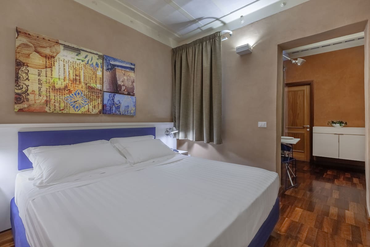 Un cuarto de hotel con una cama grande, una mesita de noche, una lámpara de pared, un cuadro, y un baño visible a través de una puerta abierta.