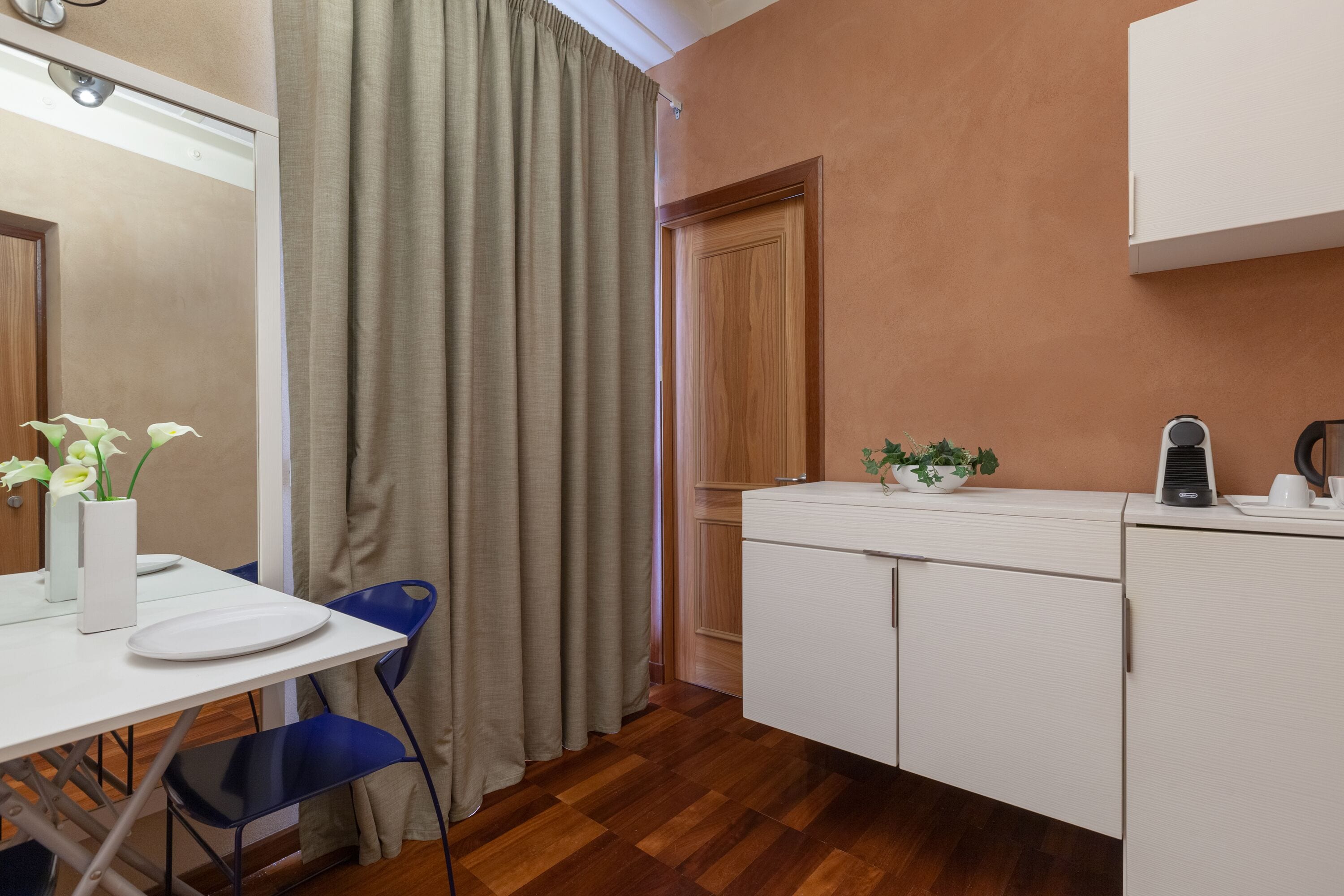 Una stanza compatta con pavimento in legno, una scrivania bianca con un vaso di fiori, una sedia blu, un armadietto bianco e una porta che conduce a un'altra stanza.