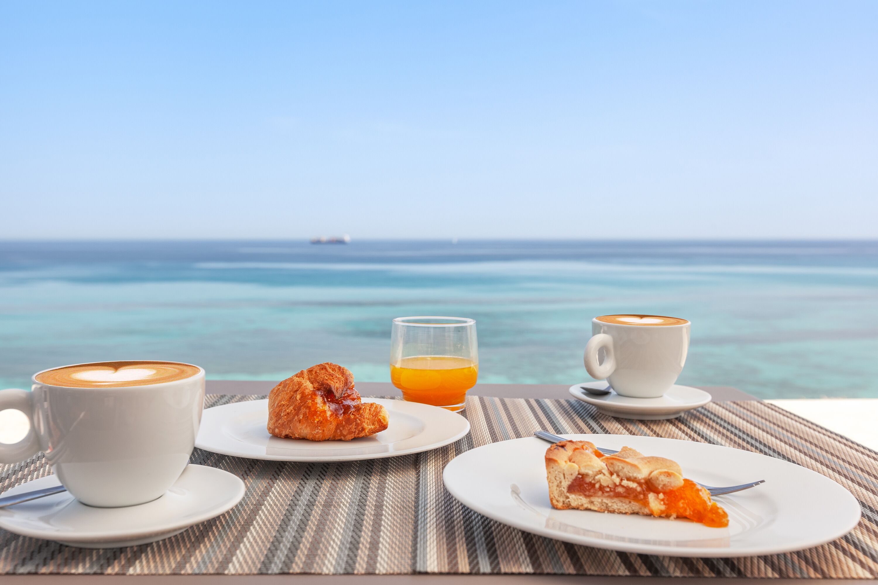 A table with two cups of coffee, a glass of orange juice, and a croissant and pizza slice on plates, overlooking a beach with clear water and a distant ship.