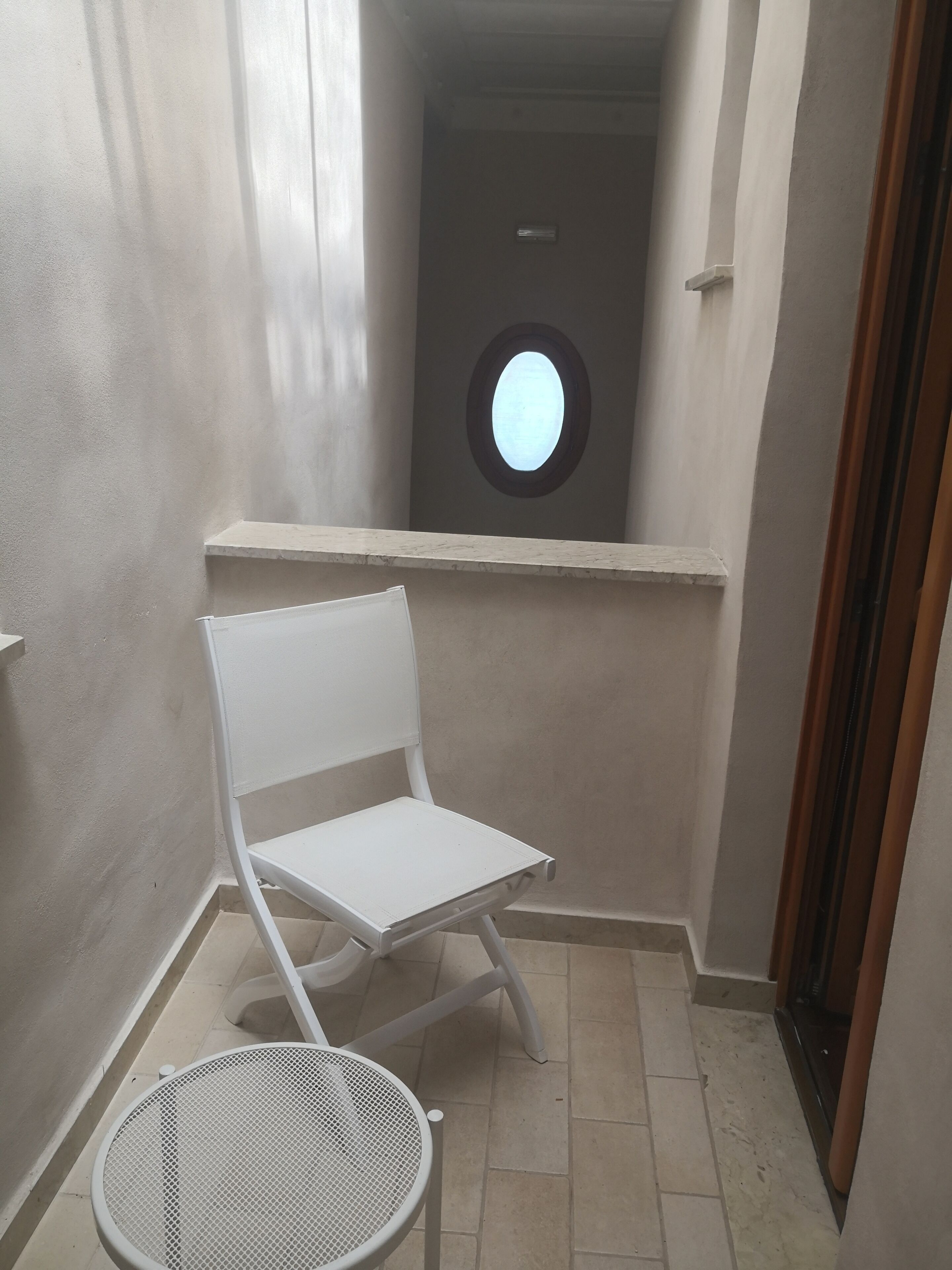 Un pasillo estrecho con una silla blanca y una mesita, un ventanal redondo y una puerta al final.