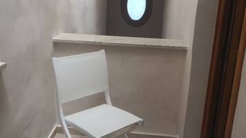 Un pasillo estrecho con una silla blanca y una mesita, un ventanal redondo y una puerta al final.