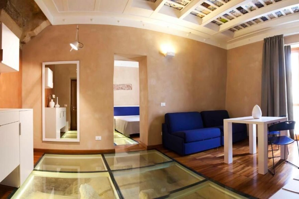 A modern living room with a glass coffee table, a blue sofa, a white table, and a mirror.