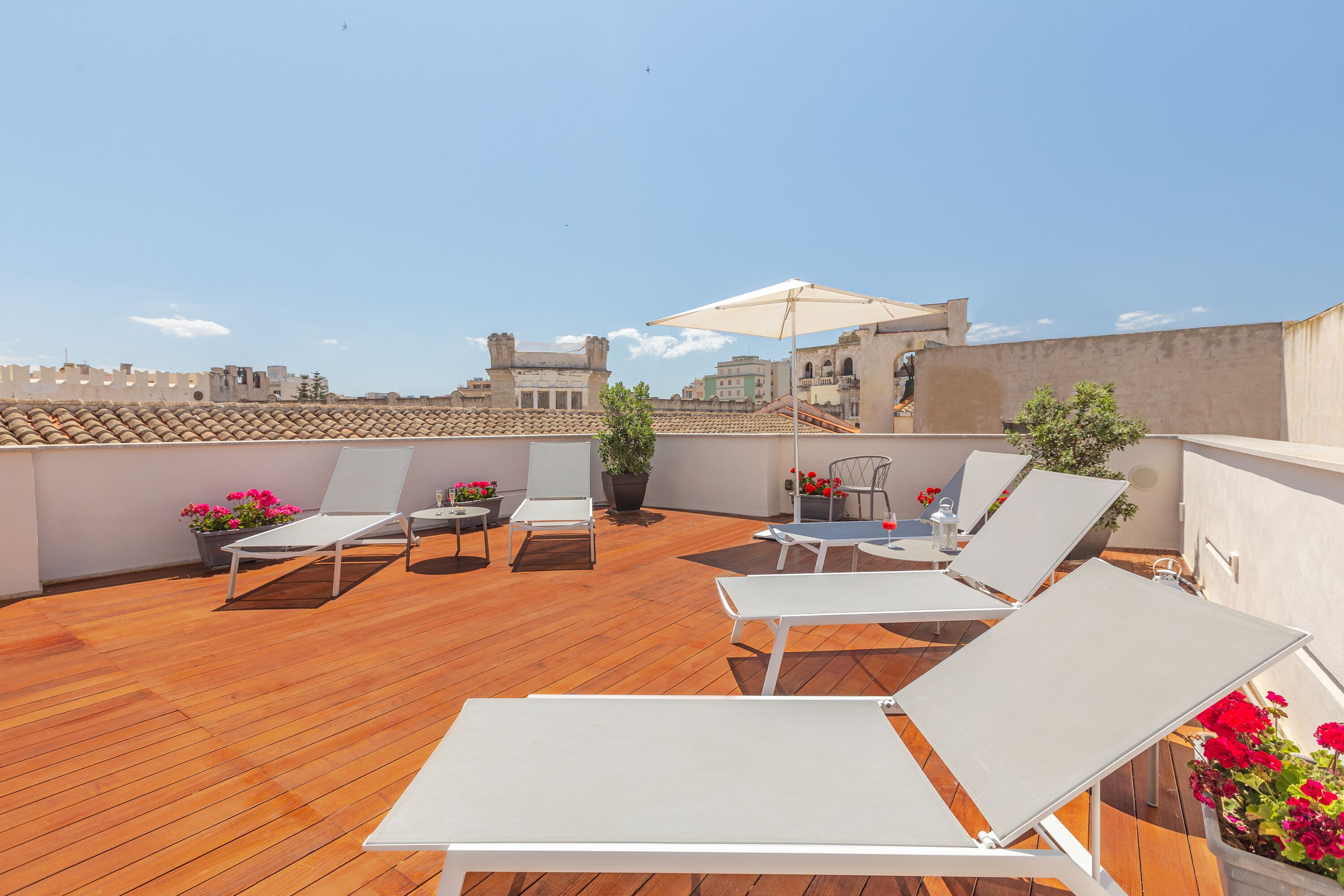 A rooftop terrace with wooden flooring, white lounge chairs, a table, and potted plants with red flowers.
