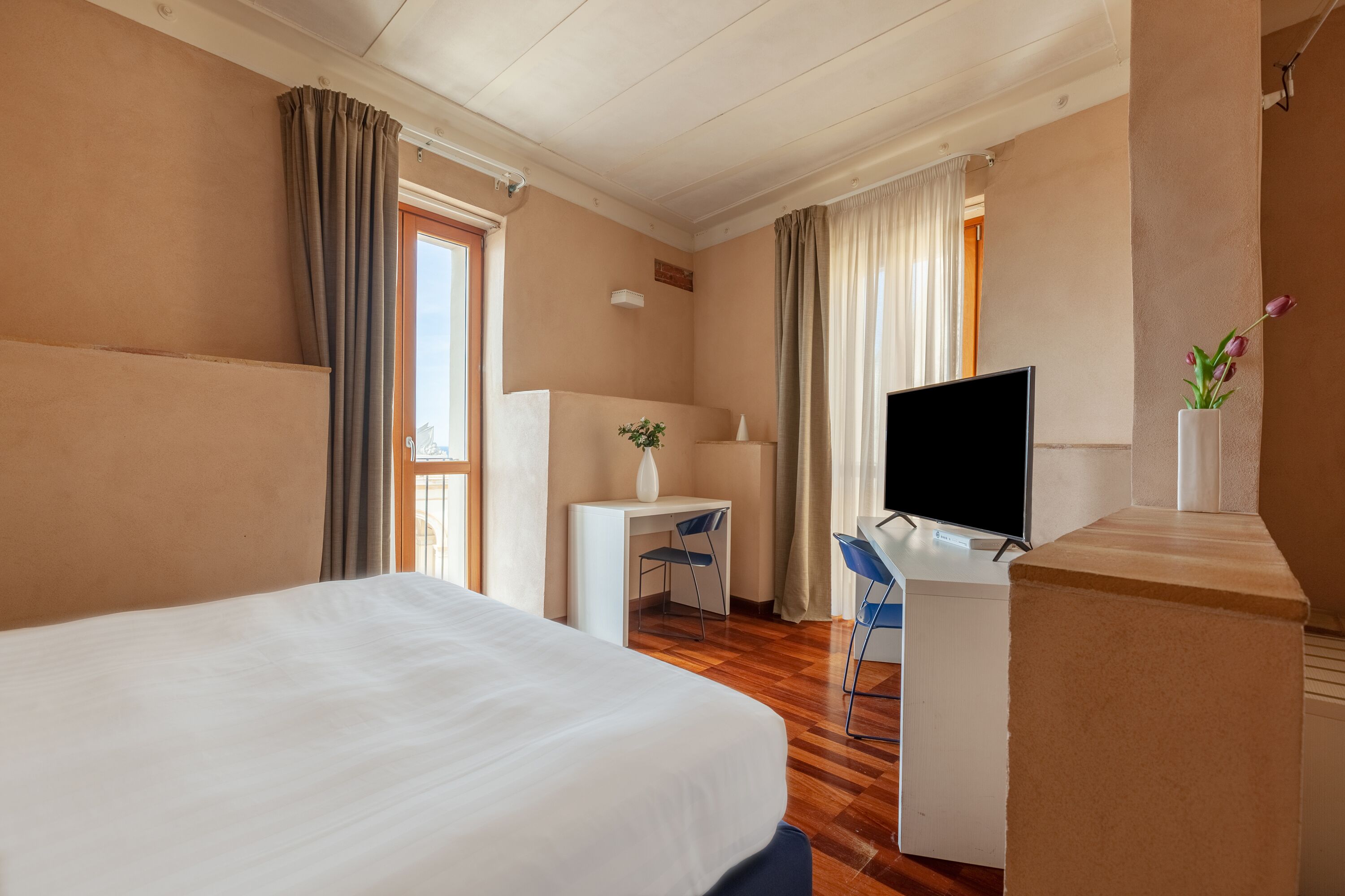 A hotel room with a large bed, a desk with a chair, a television, and a small table with a vase of flowers.