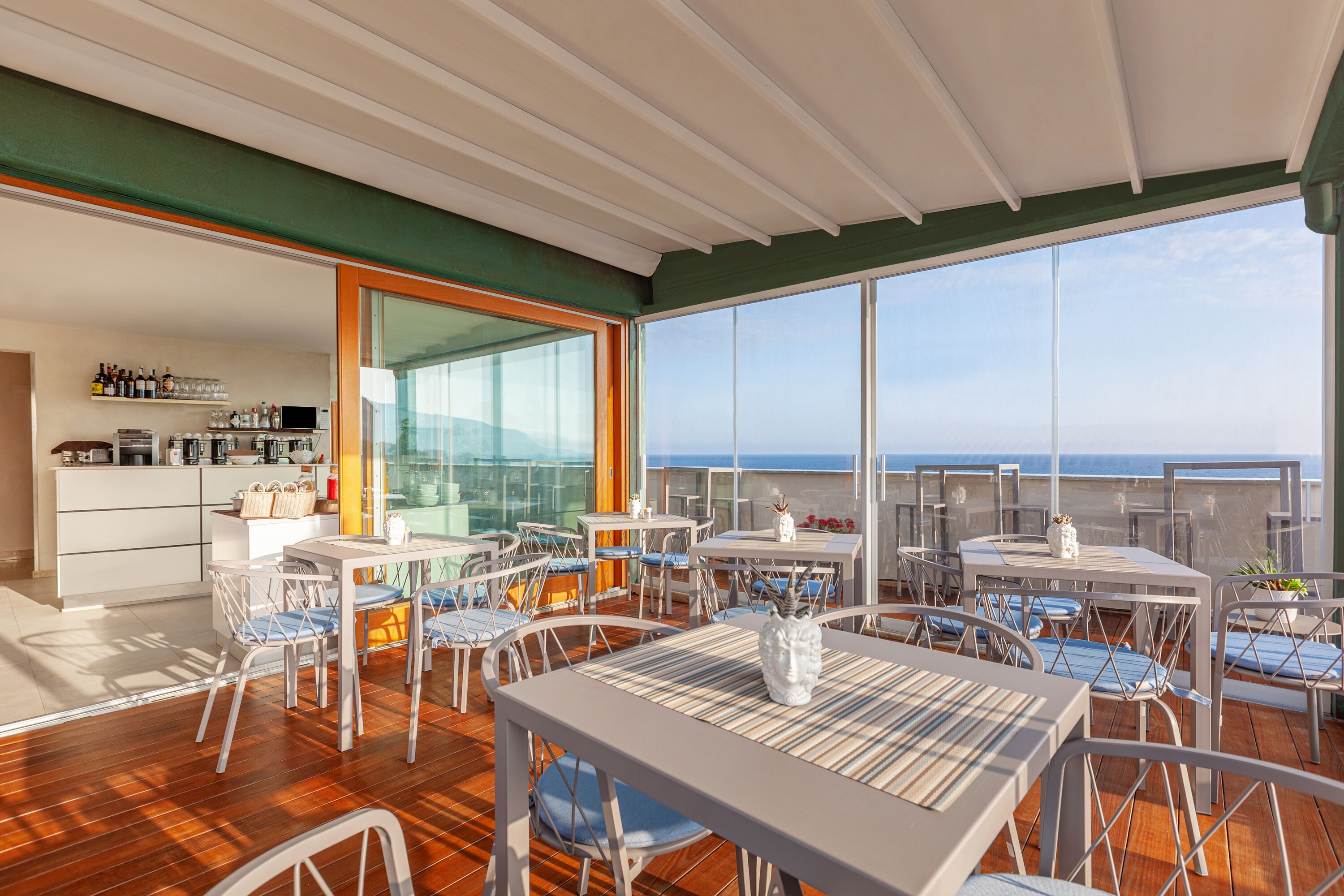 Un'ampia terrazza esterna con diversi tavoli e sedie, un'area bar con scaffali pieni di bottiglie e un'ampia vetrata che si affaccia sull'oceano.