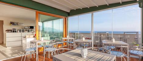 A spacious outdoor deck with multiple tables and chairs, a bar area with shelves of bottles, and a large glass window offering a view of the ocean.