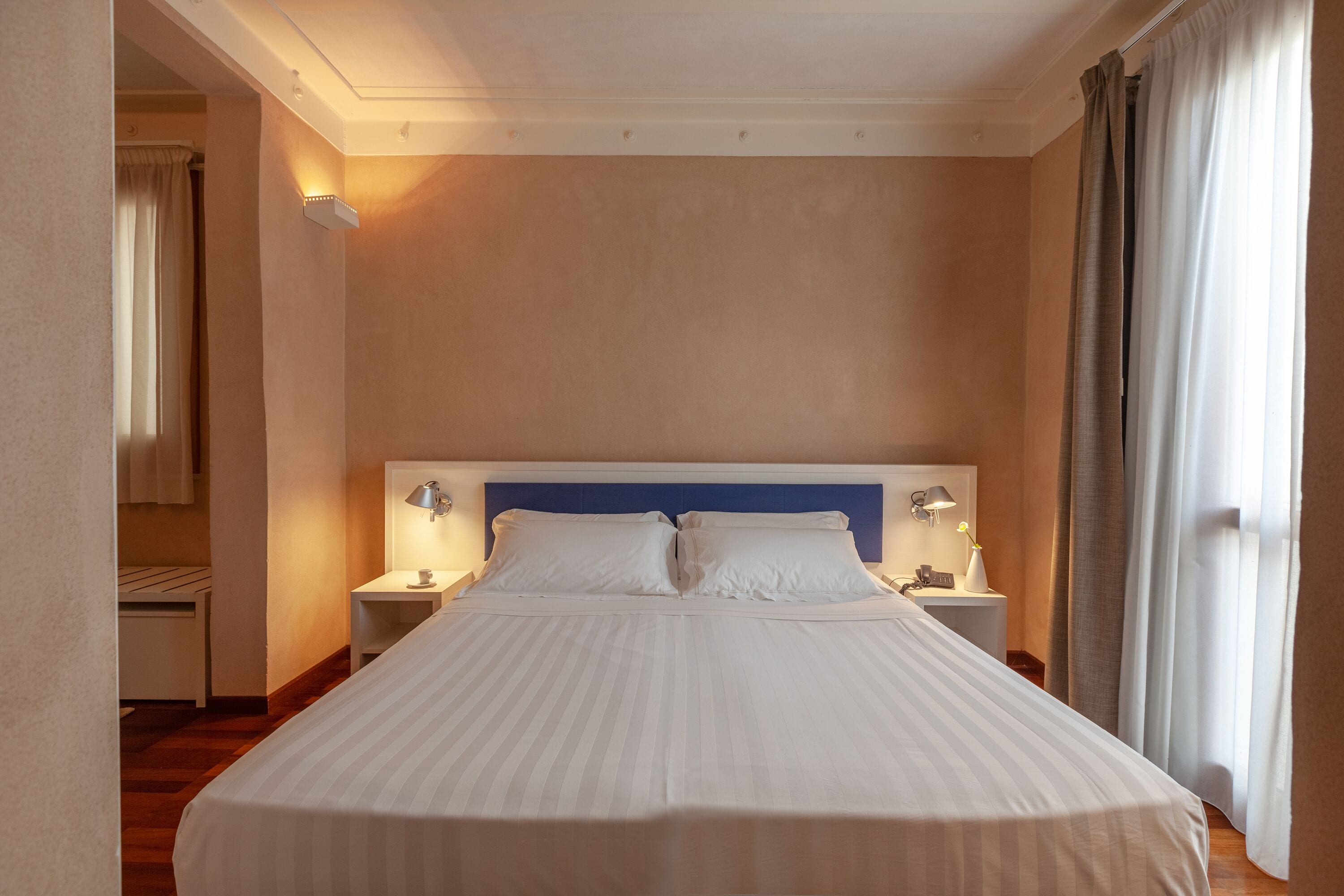 A hotel room with a large bed, two bedside tables with lamps, a headboard, and a window with curtains.