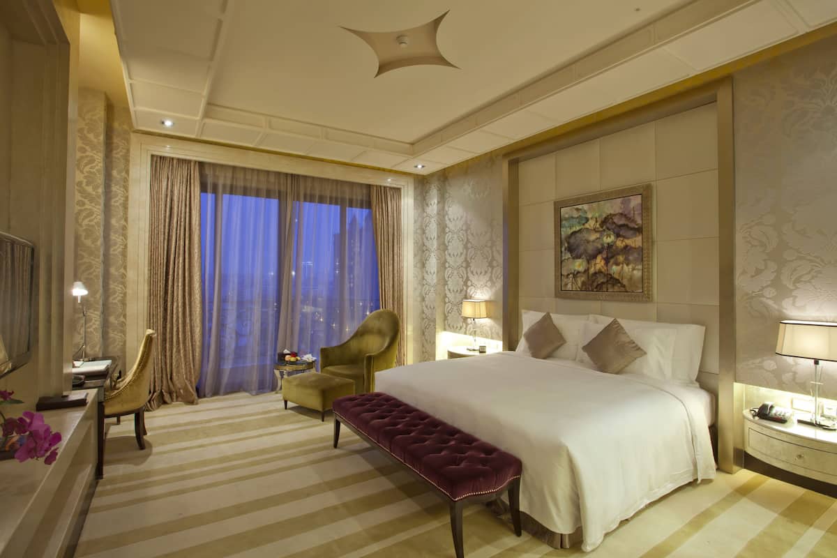 A hotel room with a large bed, a sitting area with a bench, a desk with a lamp, and a large window with curtains.