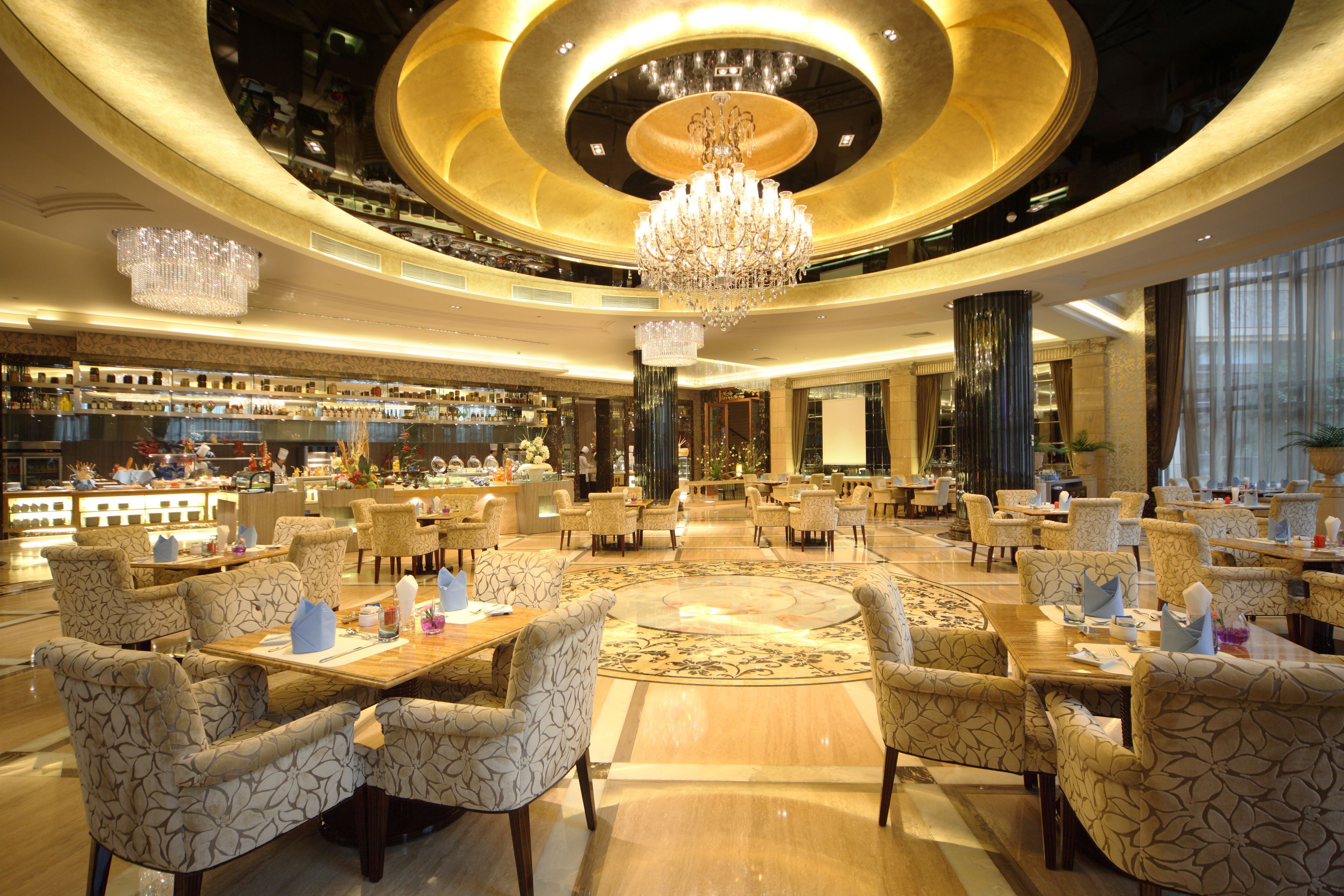 A spacious hotel lobby with a central chandelier, multiple seating areas, and a bar in the background.